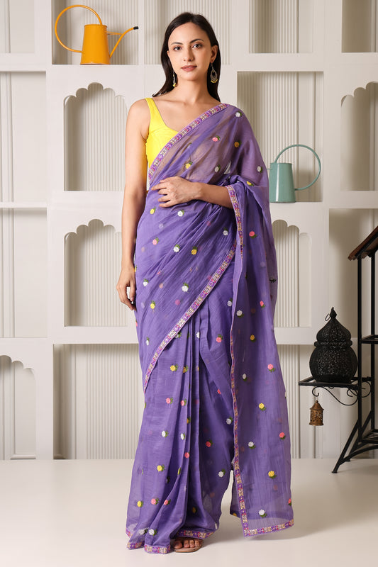 A woman models a purple saree with colorful embroidered dots and a yellow sleeveless blouse, standing in front of a white decorative wall with shelves.
