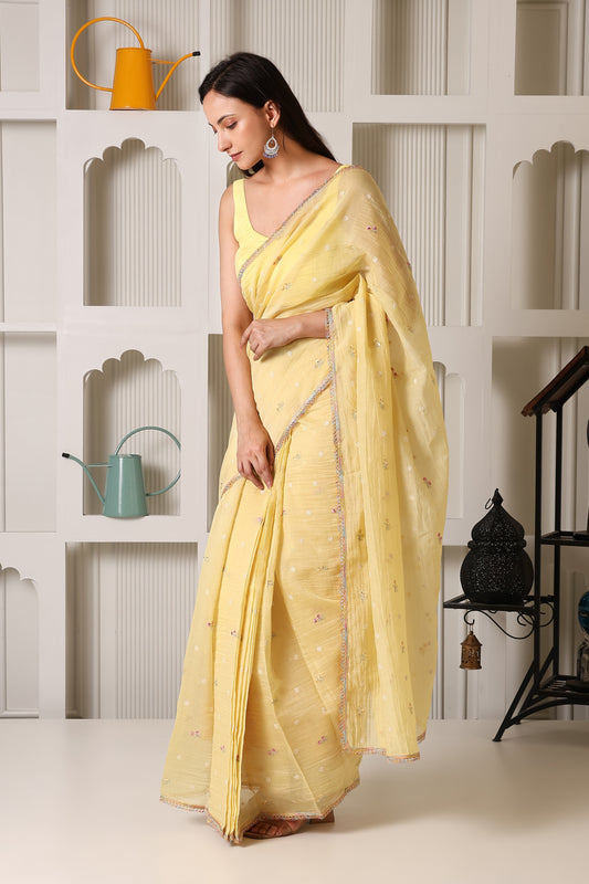 A woman wearing a light yellow saree with a matching blouse, standing in front of a white decorative wall with recessed shelves and small decorative items.