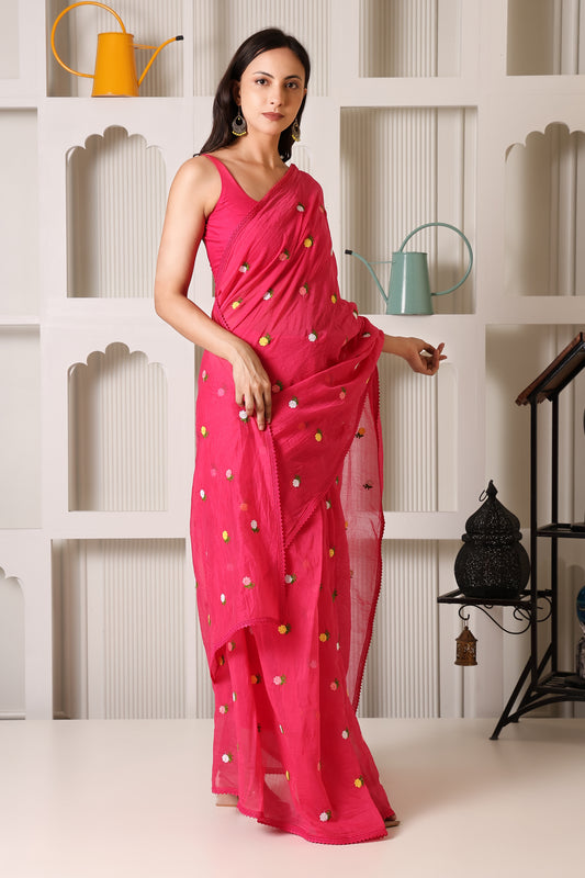 A woman models a bright pink sleeveless saree with small multicolored embroidered dots, standing in front of a white decorative wall with shelves holding colorful watering cans.