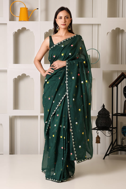 A woman models a dark green saree with small multicolored floral motifs and a silver border, standing in front of a white decorative wall.