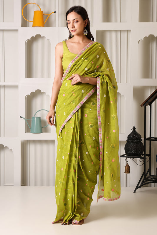 A woman models a bright lime green saree with delicate floral prints and a pink border, paired with a matching sleeveless blouse, set against a white decorative wall with shelves.