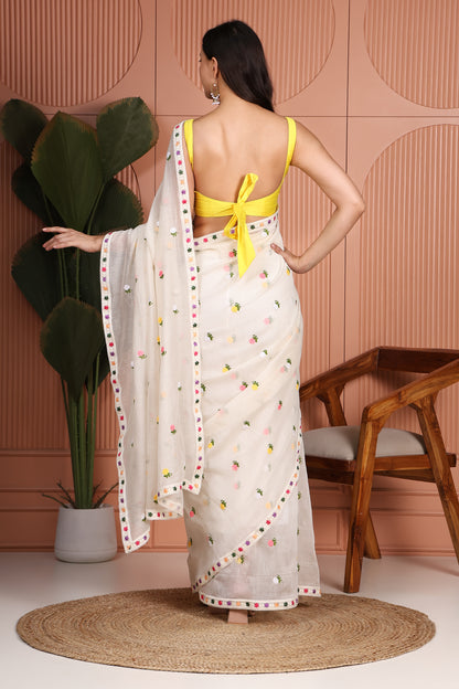A woman stands with her back to the camera wearing a white saree with colorful floral embroidery and a bright yellow backless blouse. The setting features a pink textured wall, a potted plant, a wooden chair, and a round jute rug.