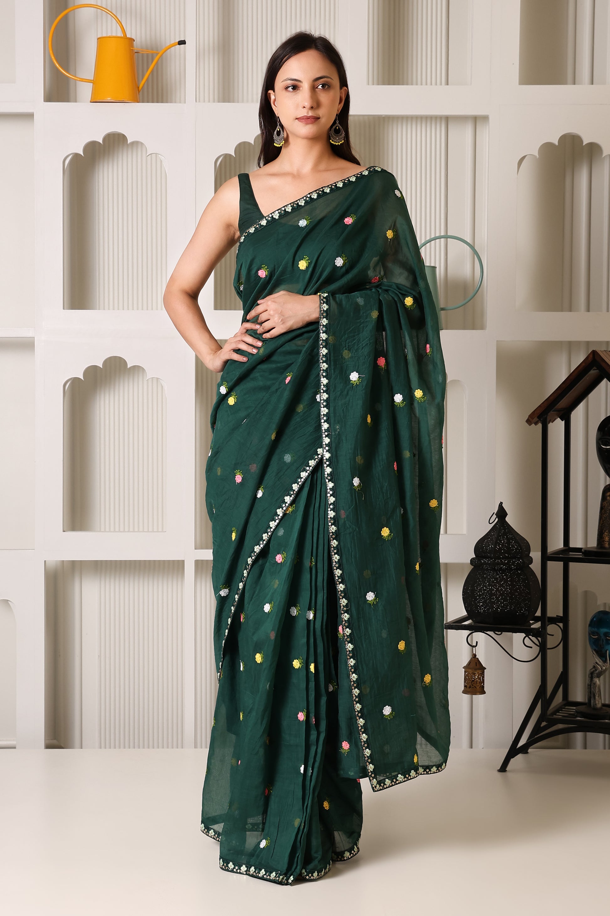 A woman models a dark green saree with small multicolored floral motifs and a silver border, standing in front of a white decorative wall.